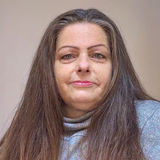 Gina, a middle-aged woman with long brown hair and a neutral expression, wears a gray turtleneck sweater as she poses against a plain background, reflecting her quiet strength in helping others when they needed help most.