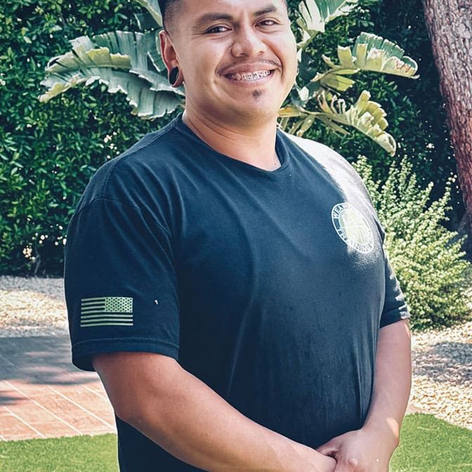 A person stands outdoors on grass, smiling at the camera—a moment reflecting Jose’s life and the gift of desperation. They wear a black T-shirt with an American flag patch on the sleeve and have short dark hair. Green plants are in the background.