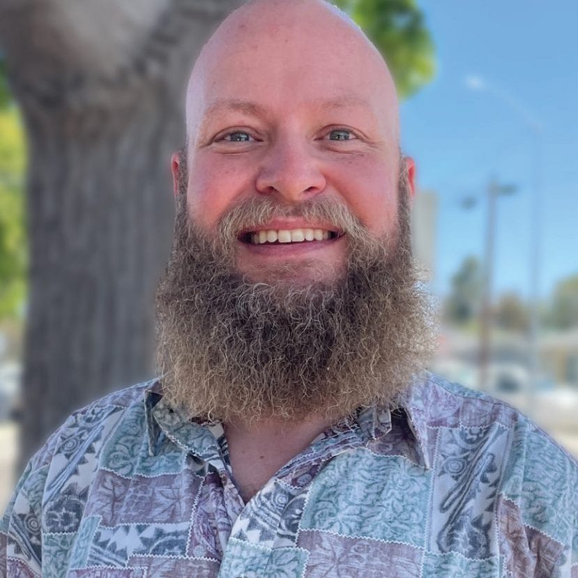 Smiling man with a beard outdoors near a tree and blue sky.