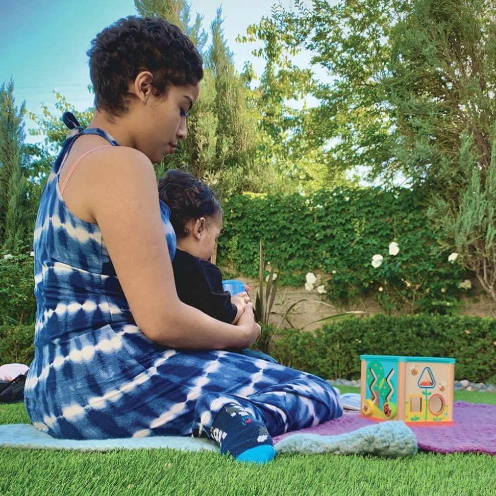 A person in a tie-dye outfit sits on the grass with a child on their lap, next to a colorful activity block, surrounded by greenery. Director Josie Casarrubias enjoys this serene moment at RMA San Fernando Valley.