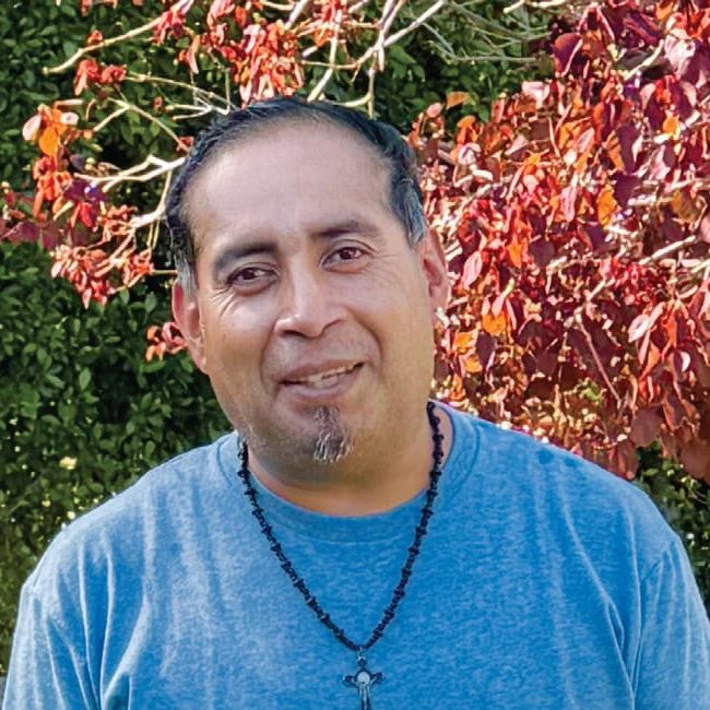 A compassionate man smiling outdoors at San Fernando Rescue Mission in front of colorful foliage.