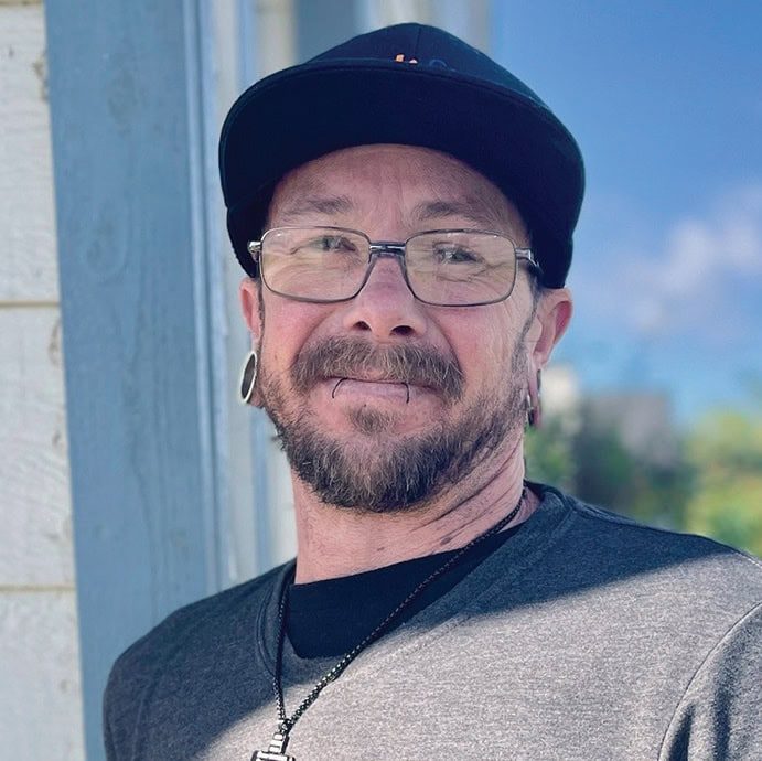 A smiling man with glasses and a black cap outdoors at San Fernando Rescue Mission.